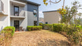 Modern two-story residential building exterior with a dry grassy yard, trimmed hedges, trees, and a clear blue sky.