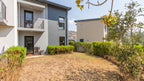 Modern two-story residential building exterior with a dry grassy yard, trimmed hedges, trees, and a clear blue sky.