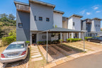 Modern multi-story townhouse villas with gray and white facades, featuring paved front yards with a black carport, parked cars, and a clear blue sky.